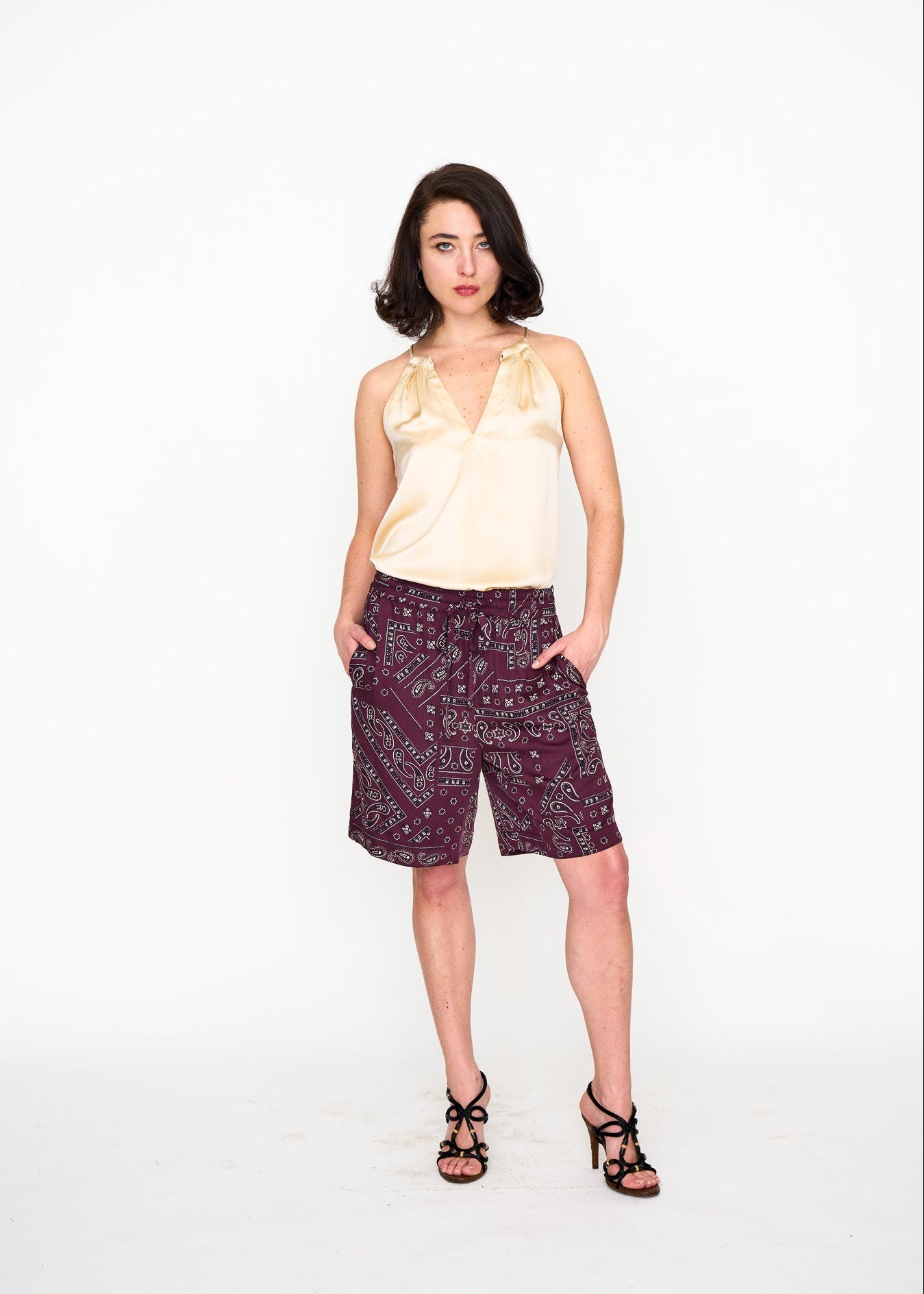 Woman wearing a light-colored silk halter top and patterned Bermuda shorts on a white background